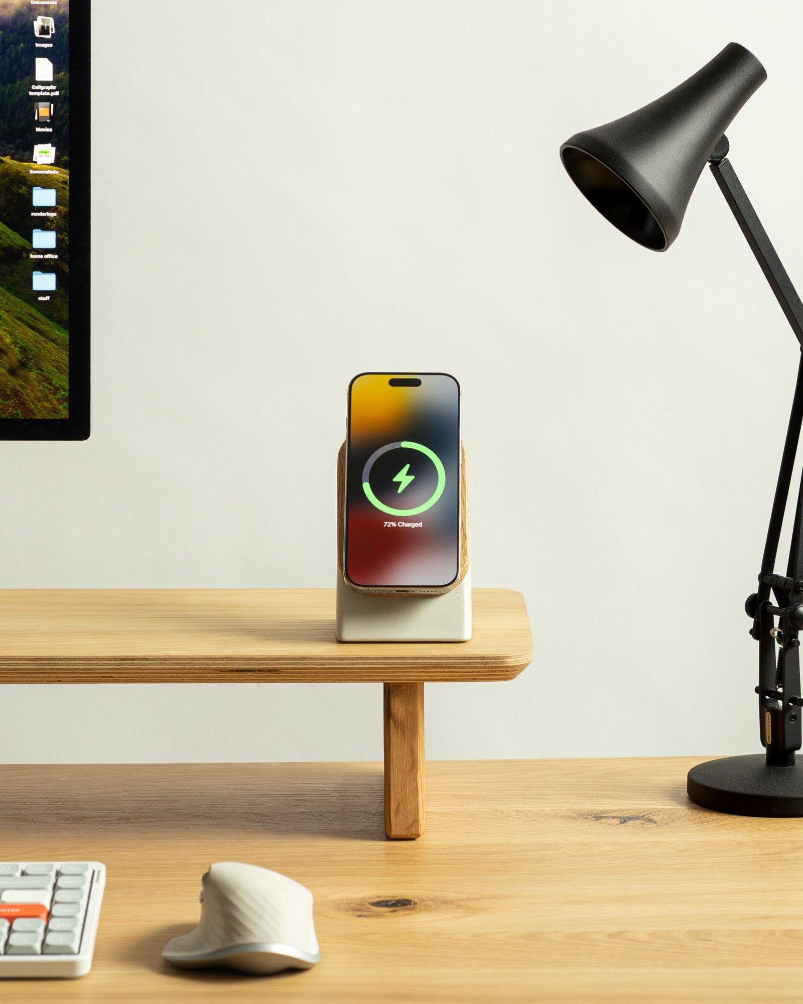 Smartphone on a stand with charging icon on a desk next to a computer monitor and lamp.