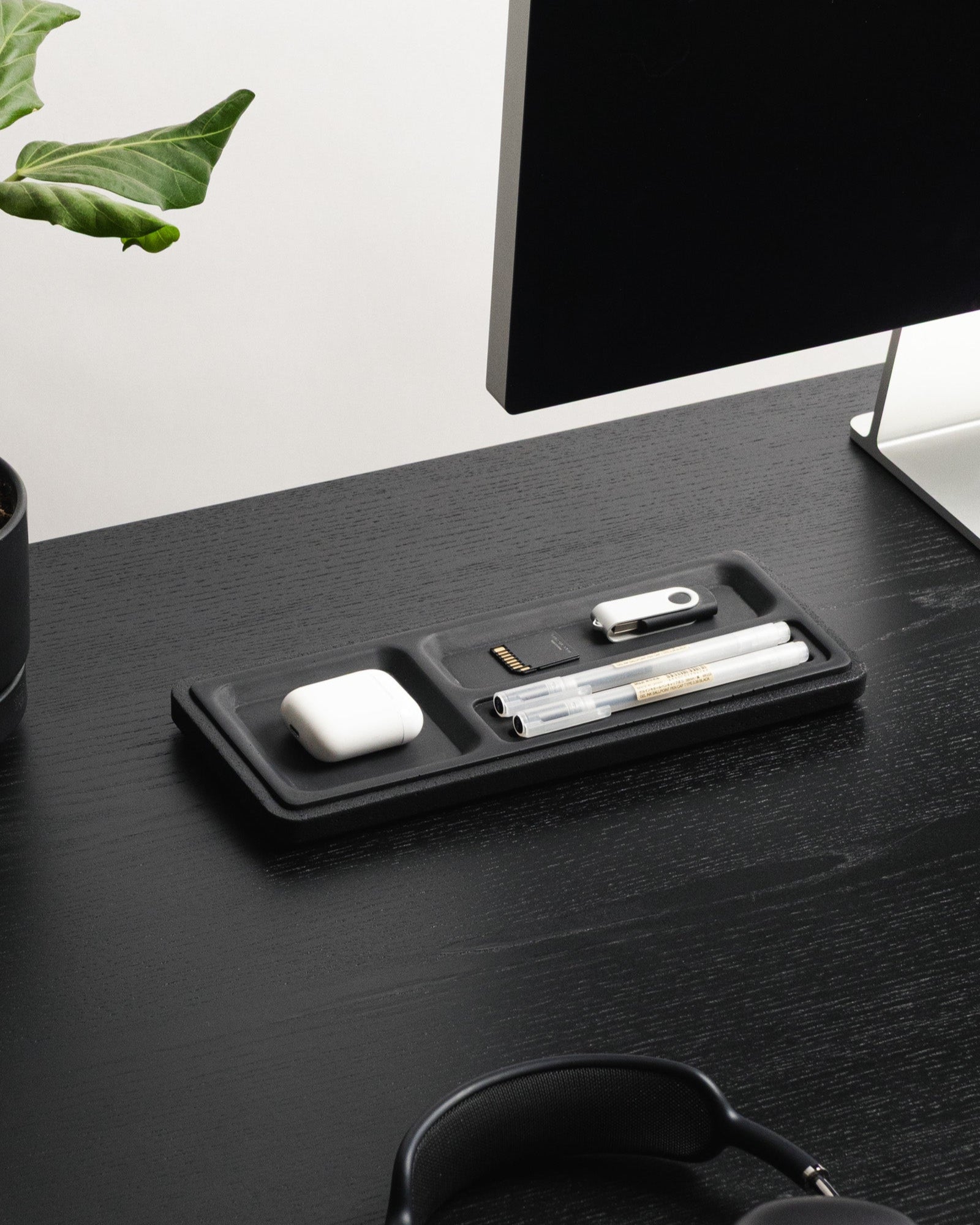 Black desk organizer with electronic devices on a dark surface with a plant in the corner.