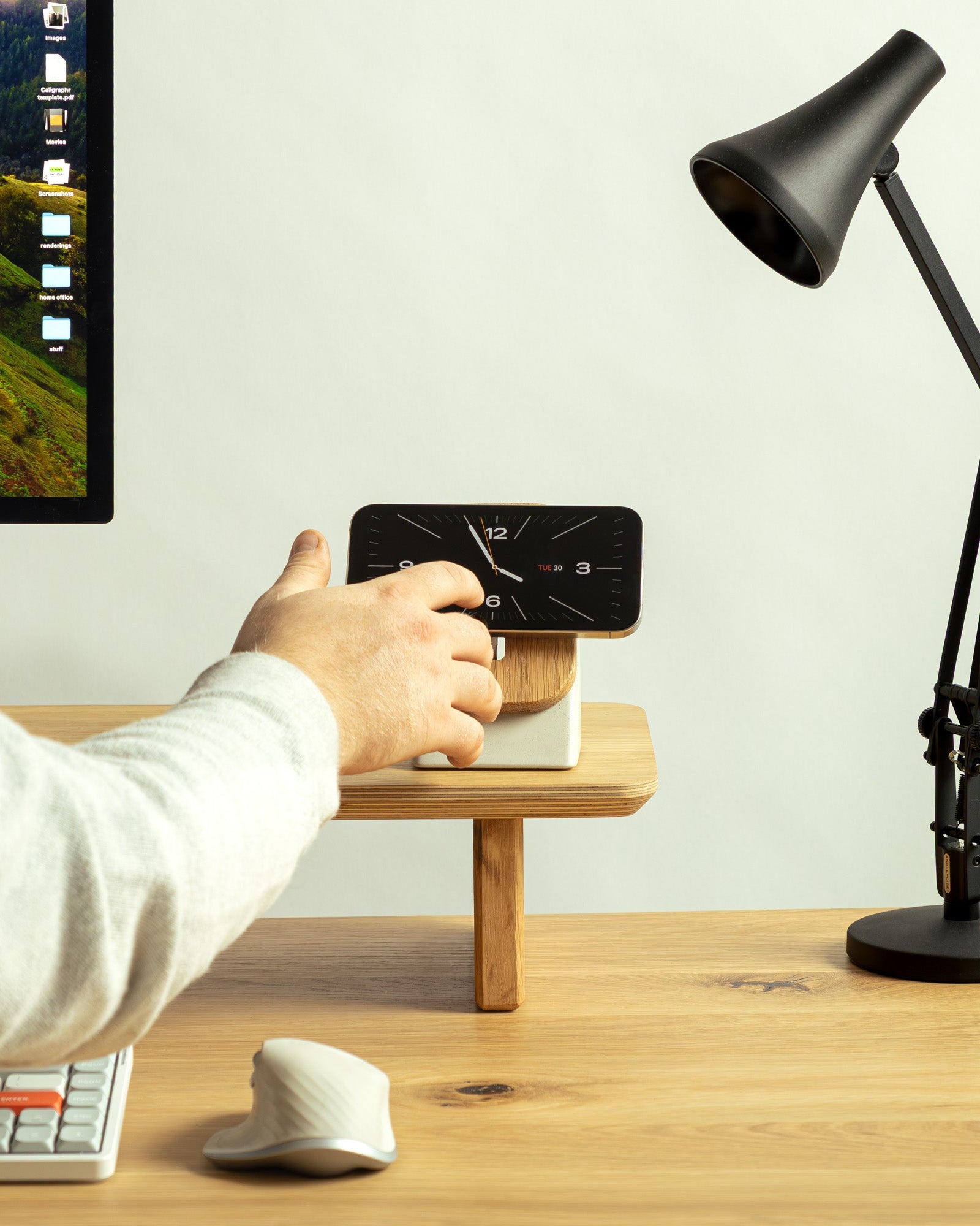 Person using a smartphone with a dock on a wooden desk next to a lamp and computer monitor.