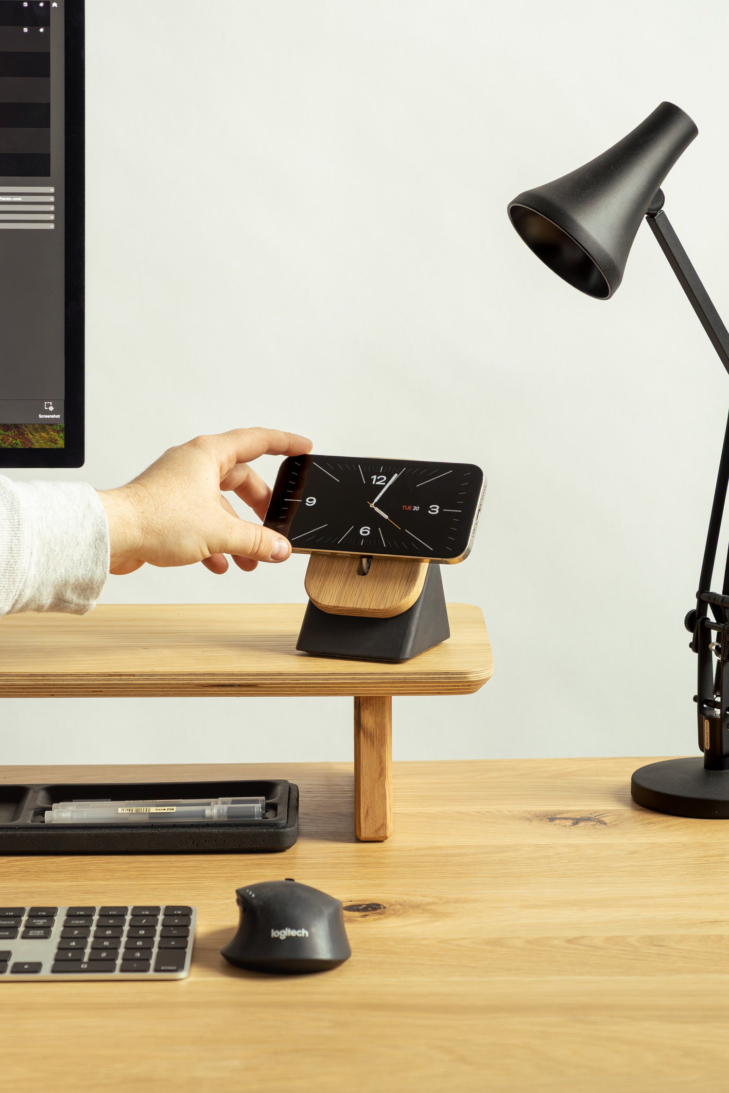 Person using a smartphone with a stand on a desk next to a computer monitor and lamp.