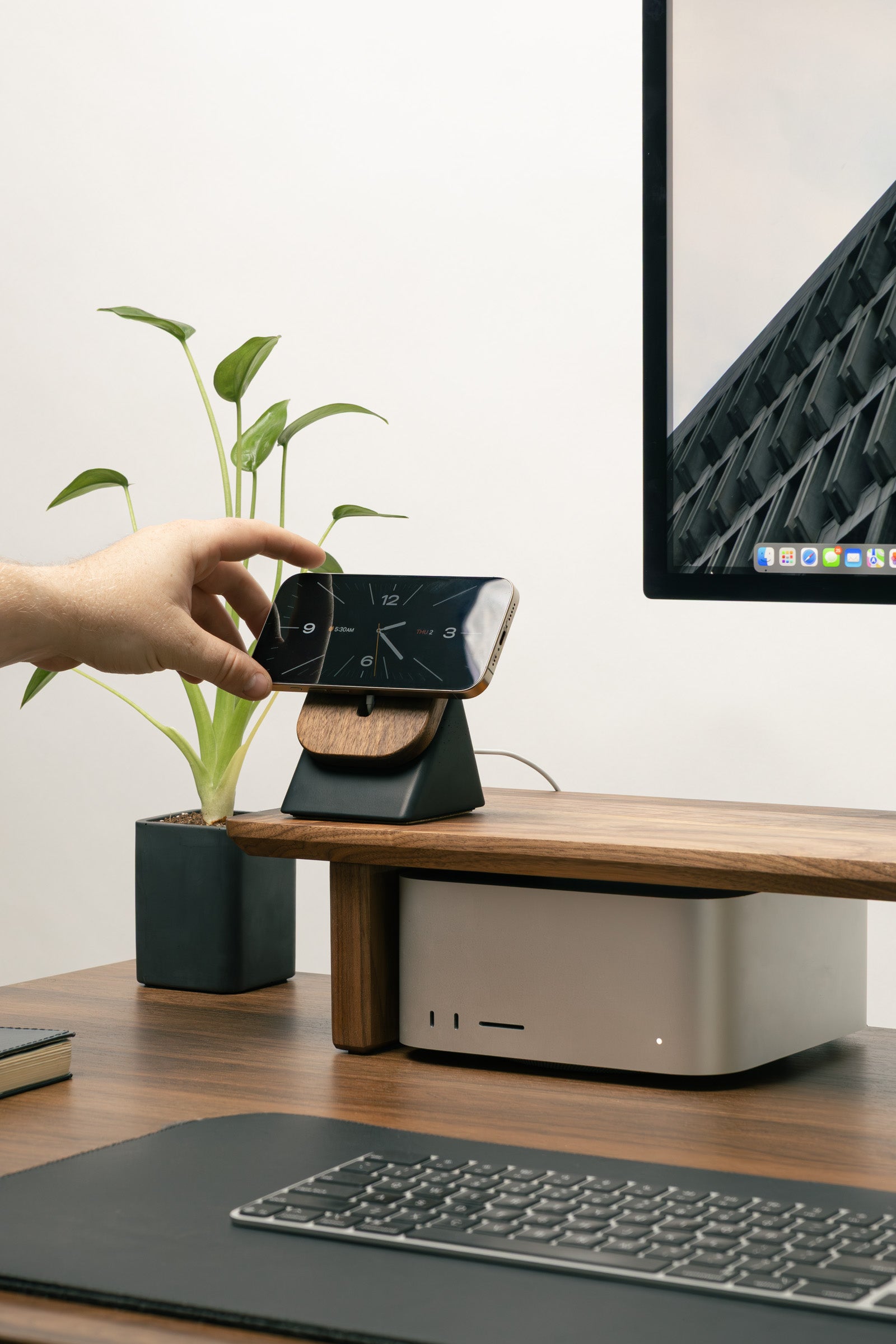 Hand holding a smartphone with a wireless charging stand on a desk with a computer monitor and keyboard.