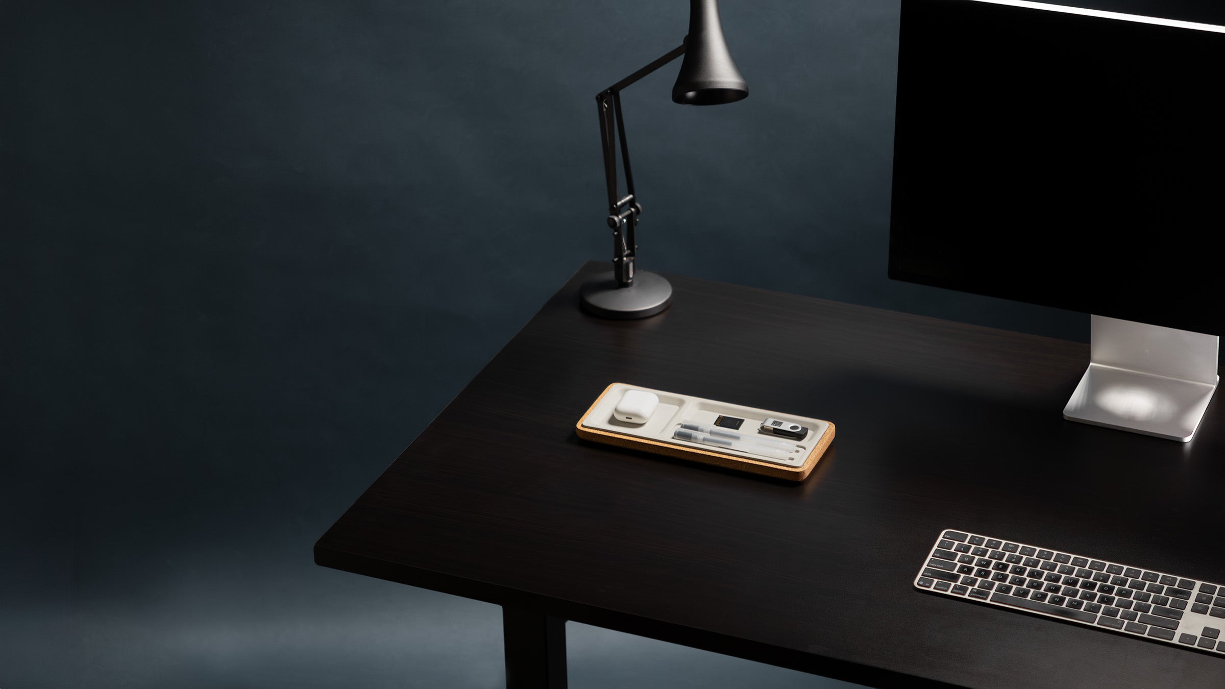 Dark office desk with computer monitor, keyboard, and lamp against a dark background